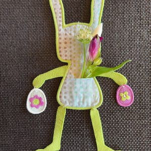 Puede incluir: Un conejito de fieltro verde con un bolsillo que contiene un ramo de flores blancas y moradas. El conejito sostiene dos huevos de fieltro rosa y blanco con flores.