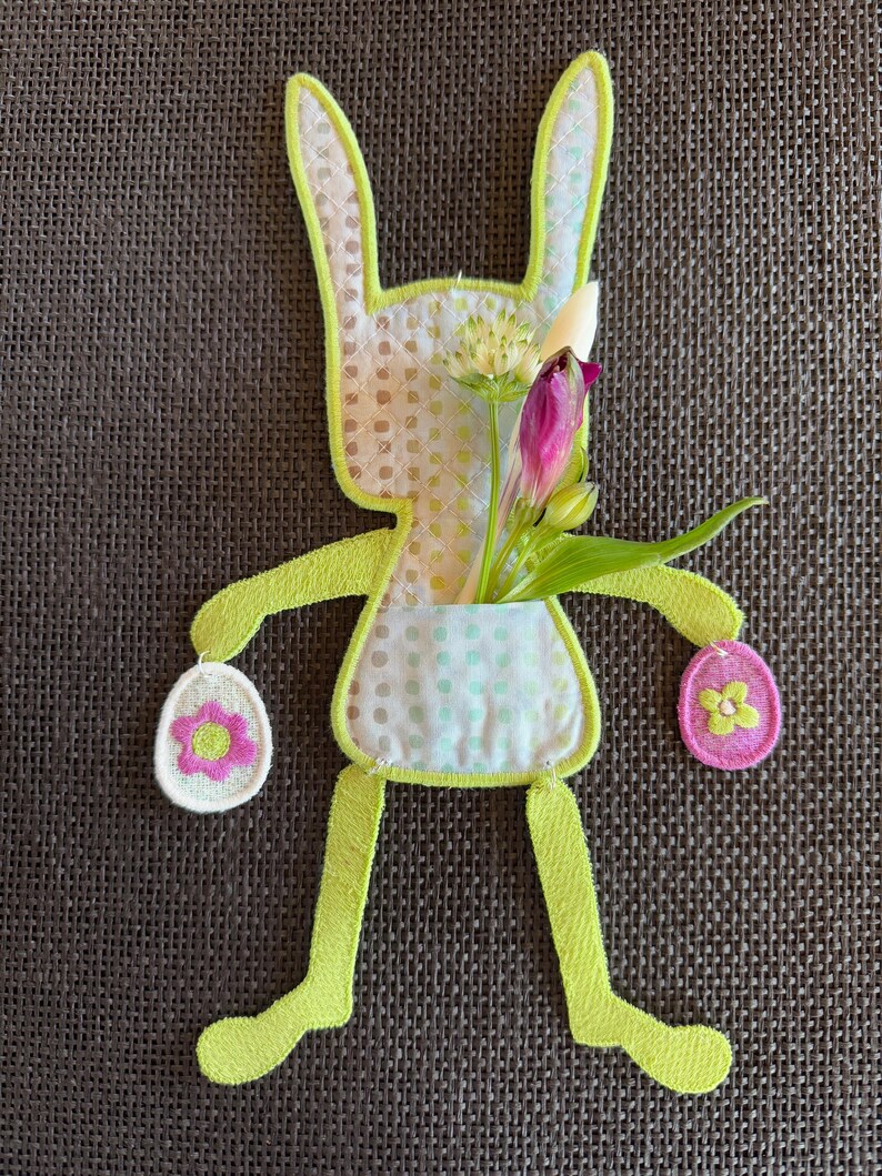 Puede incluir: Un conejito de fieltro verde con un bolsillo que contiene un ramo de flores blancas y moradas. El conejito sostiene dos huevos de fieltro rosa y blanco con flores.
