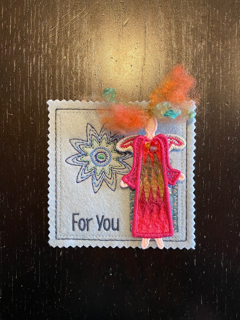Puede incluir: Una tarjeta de fieltro hecha a mano con una flor bordada y una figura de &aacute;ngel de fieltro. La tarjeta tiene el texto "Para Ti" bordado.