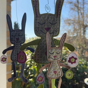 Puede incluir: Tres adornos de conejo bordados colgando de una ventana. Los conejos sostienen huevos de Pascua con flores. Los conejos son de color verde, marr&oacute;n y gris.
