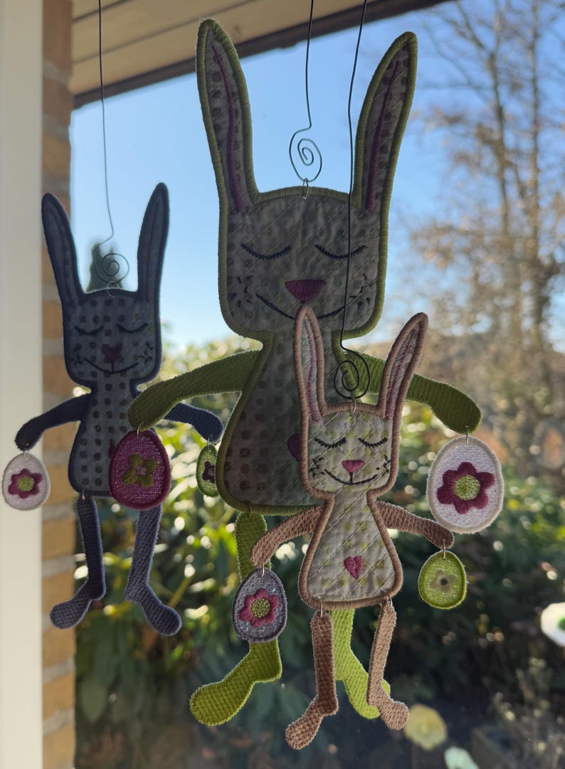 Puede incluir: Tres adornos de conejo bordados colgando de una ventana. Los conejos sostienen huevos de Pascua con flores. Los conejos son de color verde, marr&oacute;n y gris.