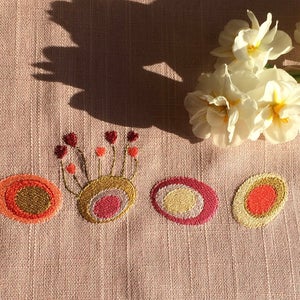 Può includere: Un tessuto di lino rosa con cerchi ricamati in tonalità di arancio, rosso, oro e crema. I cerchi sono disposti in fila e presentano un motivo floreale stilizzato al centro.