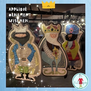 May include: Appliqué ornament of three wise men, each with a crown and detailed clothing. The ornaments are in shades of gold, blue, green, and white, with the text "APPLIQUÉ ORNAMENT WISEMEN" at the top. They are hanging from a string of lights.
