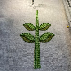 Puede incluir: Un tallo de tela verde con tres hojas de tela verde, cosidas sobre un fondo de tela beige claro.