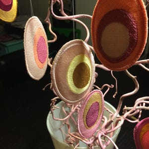 Op de afbeelding: Een mobiel gemaakt van stoffen cirkels met verschillende kleuren en patronen. De cirkels hangen aan een takachtige structuur. De kleuren omvatten roze, oranje, geel en bruin.