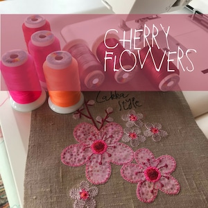 Op de afbeelding: Close-up van een naaiproject met roze en rode kersenbloesem-borduurwerk op een beige linnen stof. Vijf spoelen borduurgaren in verschillende tinten roze en oranje zijn zichtbaar op de achtergrond. De tekst "CHERRY FLOWERS Lakka Style" is zichtbaar boven de borduurwerk.