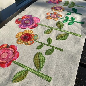 Puede incluir: Un camino de mesa de lino blanco con cinco flores bordadas de colores. Cada flor tiene un patr&oacute;n y una combinaci&oacute;n de colores diferentes, incluyendo rosa, rojo, amarillo, verde y morado. Las flores est&aacute;n dispuestas en una fila en el centro del camino de mesa.
