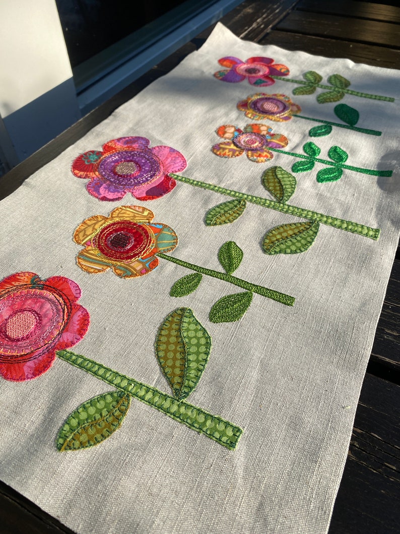 Puede incluir: Un camino de mesa de lino blanco con cinco flores bordadas de colores. Cada flor tiene un patr&oacute;n y una combinaci&oacute;n de colores diferentes, incluyendo rosa, rojo, amarillo, verde y morado. Las flores est&aacute;n dispuestas en una fila en el centro del camino de mesa.