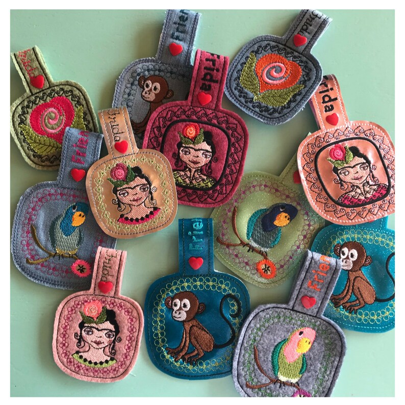 May include: A collection of 10 colorful felt keychains with embroidered designs. The designs include a monkey, a bird, a parrot, a flower, and a woman with a flower in her hair. The keychains are all attached to a loop with a heart-shaped charm that says "Frida".