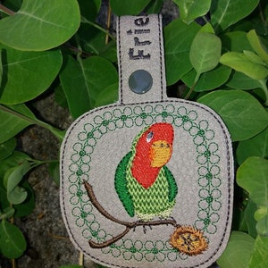 May include: A gray leather keychain with a green and red embroidered lovebird perched on a branch. The keychain has a loop for attaching keys and the word "Ellie" is embroidered on the top.