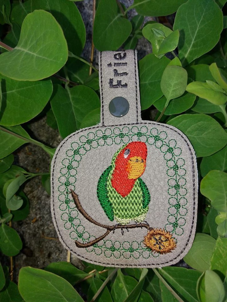 May include: A gray leather keychain with a green and red embroidered lovebird perched on a branch. The keychain has a loop for attaching keys and the word "Ellie" is embroidered on the top.