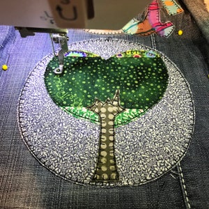 May include: A close-up of a green and brown fabric tree applique being sewn onto a denim background. The tree is surrounded by a circle of white and purple floral fabric.