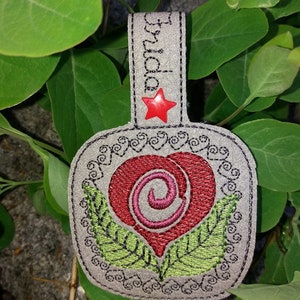 May include: A gray leather heart-shaped keychain with a red heart and green leaves embroidered on it. The keychain has a black star with a red center and the word "Grandma" embroidered on the top.