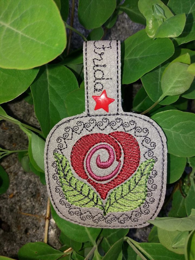 May include: A gray leather heart-shaped keychain with a red heart and green leaves embroidered on it. The keychain has a black star with a red center and the word "Grandma" embroidered on the top.