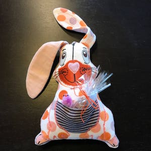 Op de afbeelding: Een roze en oranje geblokte pluchen konijn met een zakje op zijn buik. Het zakje heeft een gestreepte voering en een klein paars pluche vogeltje erin. Het konijn houdt een doorzichtig plastic zakje vast met een rood lint.