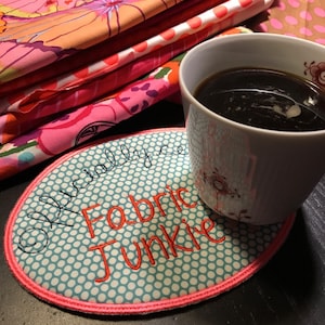 May include: An oval fabric coaster with the text "Officially Fabric Junkie" in red and black embroidery. The coaster has a light blue polka dot pattern and a pink border. A white mug with coffee sits next to the coaster, with colorful fabric in the background.