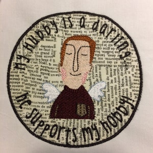 May include: A round embroidered patch with a cartoon illustration of a person with wings. The person is wearing a brown shirt and has closed eyes. The text "My hubby is a darlin'" and "He supports my sports" is visible around the person.