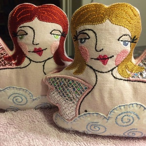 Puede incluir: Dos ángeles de tela bordados con detalles en rosa y azul. Cada ángel tiene una nube con un diseño de espiral azul. Un ángel tiene el pelo rojo y los ojos verdes, el otro tiene el pelo rubio y los ojos azules.