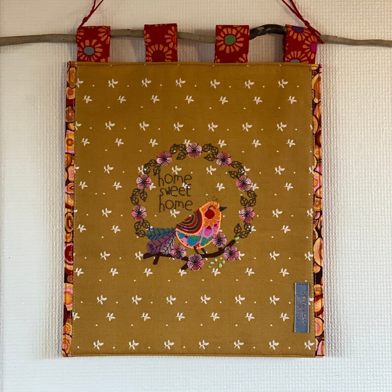 May include: A brown fabric wall hanging with a colorful embroidered bird and the text "home sweet home". The hanging has a red, white, and yellow floral border.