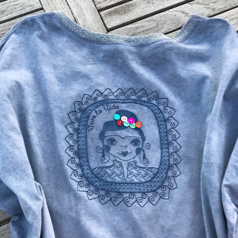 Puede incluir: Una camiseta azul claro con un dise&ntilde;o bordado de una mujer con una corona de botones coloridos. El dise&ntilde;o est&aacute; enmarcado con un borde decorativo y el texto "Viva la Vida" est&aacute; escrito sobre la cabeza de la mujer.