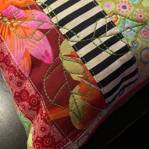 May include: A colorful patchwork pillow with a floral design and green embroidered leaves. The pillow is made of different fabrics, including red, pink, orange, black, and white.