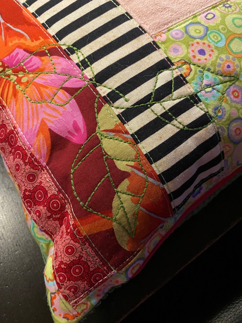 May include: A colorful patchwork pillow with a floral design and green embroidered leaves. The pillow is made of different fabrics, including red, pink, orange, black, and white.