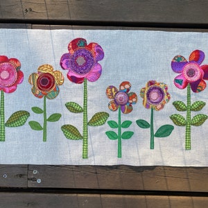 Puede incluir: Un panel de tela blanco con seis flores de tela coloridas aplicadas sobre &eacute;l. Las flores son en tonos de rosa, morado, rojo y amarillo. Las flores son todas de diferentes formas y tama&ntilde;os. El panel de tela es perfecto para un proyecto de manualidades o decoraci&oacute;n del hogar.