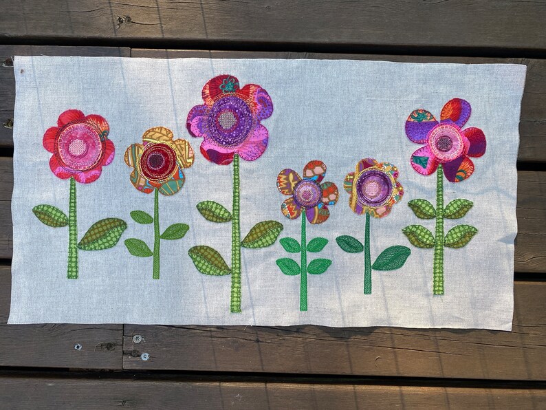 Puede incluir: Un panel de tela blanco con seis flores de tela coloridas aplicadas sobre &eacute;l. Las flores son en tonos de rosa, morado, rojo y amarillo. Las flores son todas de diferentes formas y tama&ntilde;os. El panel de tela es perfecto para un proyecto de manualidades o decoraci&oacute;n del hogar.