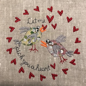 以下が含まれることがあります： 赤いハートの輪で囲まれた、くちばしを開けて大きな目をした鳥が2羽描かれた刺繍作品。鳥の下には「Let me stitch you a heart」という文字が刺繍されている。