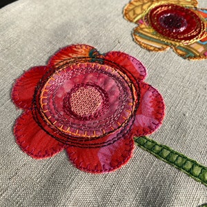 Puede incluir: Primer plano de una flor de tela roja, rosa y naranja con un tallo verde. La flor est&aacute; cosida con m&uacute;ltiples capas de tela e hilo, creando un aspecto texturizado y dimensional.