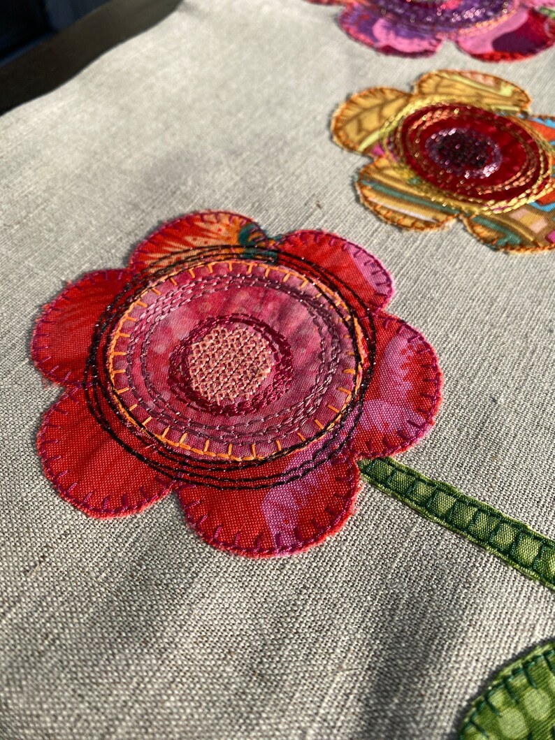 Puede incluir: Primer plano de una flor de tela roja, rosa y naranja con un tallo verde. La flor est&aacute; cosida con m&uacute;ltiples capas de tela e hilo, creando un aspecto texturizado y dimensional.