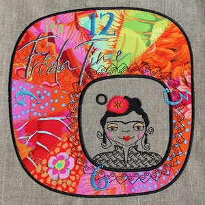 Op de afbeelding: Een kleurrijke geborduurde klok met een portret van Frida Kahlo in het midden. De wijzerplaat van de klok is omgeven door een patchwork van stof in tinten rood, oranje, roze en groen. De cijfers 12 en 6 zijn zichtbaar op de wijzerplaat van de klok. De tekst "It's Frida Time" is geborduurd bovenaan de klok.