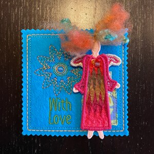 Puede incluir: Una tarjeta de fieltro azul con un dise&ntilde;o de flor cosido y las palabras "With Love" en hilo verde. Una peque&ntilde;a mu&ntilde;eca de fieltro con cabello rojo y un vestido rosa est&aacute; unida a la tarjeta.