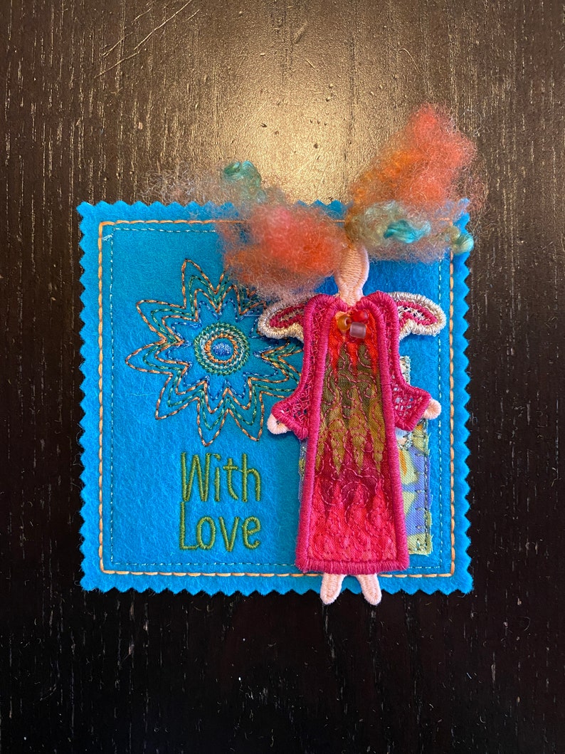 Puede incluir: Una tarjeta de fieltro azul con un dise&ntilde;o de flor cosido y las palabras "With Love" en hilo verde. Una peque&ntilde;a mu&ntilde;eca de fieltro con cabello rojo y un vestido rosa est&aacute; unida a la tarjeta.
