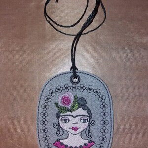 May include: A gray felt oval pendant with a black cord. The pendant features an embroidered image of Frida Kahlo with a pink flower in her hair and the word "Frida" below the image.