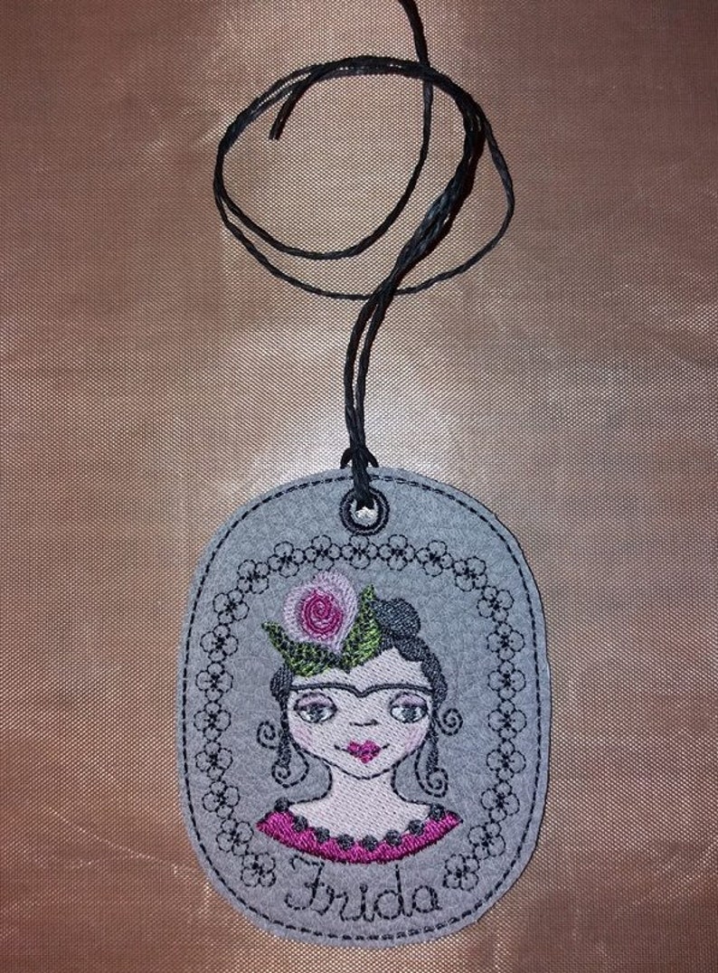 May include: A gray felt oval pendant with a black cord. The pendant features an embroidered image of Frida Kahlo with a pink flower in her hair and the word "Frida" below the image.