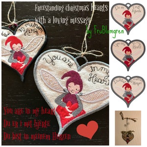 Könnte beinhalten: Drei herzförmige Ornamente mit gestickten Feen, die Herzen halten. Die Ornamente sind mit dem Text "You are in my heart", "Du er i mit hjerte" und "Du bist in meinem Herzen" verziert.