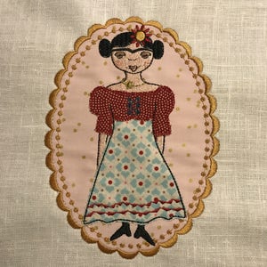 May include: Embroidered image of a woman with dark hair and a red and blue dress, wearing a flower in her hair. The image is surrounded by a gold oval frame with a scalloped edge.