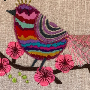 May include: Embroidered design of a colorful bird with a crown perched on a branch with pink flowers. The bird has a striped body with a purple, pink, red, orange, yellow, green, and blue pattern.