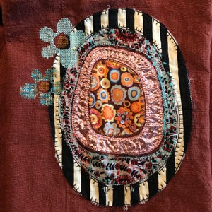 Può includere: Un'opera d'arte tessile su sfondo marrone. Presenta un design ovale con strati di tessuto concentrici. Il centro mostra un motivo floreale arancione e blu. L'ovale è bordato da strisce bianche e nere e fiori ricamati a punto croce.