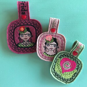 May include: Three felt keychains with embroidered designs. The first keychain is pink and features a portrait of Frida Kahlo with the word "Frida" on the top. The second keychain is pink and features a portrait of Frida Kahlo with the word "Frida" on the top. The third keychain is green and features a pink heart with a swirl design.