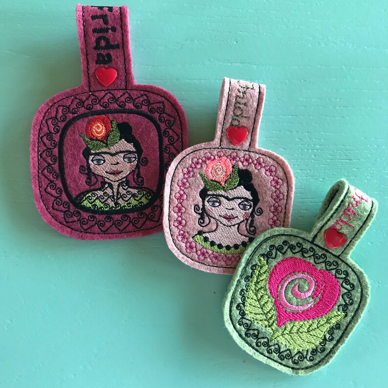 May include: Three felt keychains with embroidered designs. The first keychain is pink and features a portrait of Frida Kahlo with the word "Frida" on the top. The second keychain is pink and features a portrait of Frida Kahlo with the word "Frida" on the top. The third keychain is green and features a pink heart with a swirl design.