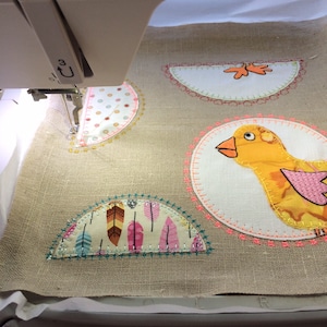 Könnte beinhalten: Nahaufnahme einer Nähmaschine, die ein Stoffprojekt näht. Das Projekt zeigt einen gelben Vogel, ein Design mit rosa und braunen Federn und einen weißen Halbkreis mit einem roten Blatt.