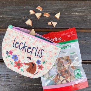 Könnte beinhalten: Eine rosa-weiße Stofftasche mit dem aufgestickten Wort "leckerlis". Die Tasche ist offen und eine Tüte Hundefutter ist sichtbar. Die Tüte ist rot-weiß und hat den Text "chrisco" und "100 ge" drauf. Die Leckerlis sind dreieckig und braun.