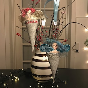 Könnte beinhalten: Drei Filz-Engel-Anhänger mit gestickten Gesichtern und Flügeln. Die Engel hängen an einem Zweig mit roten Beeren und befinden sich in einer Vase mit weißen und goldenen Streifen.