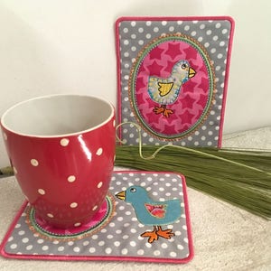 May include: Two gray and pink fabric coasters with a white polka dot pattern. One coaster features a pink oval with a yellow bird and stars. The other coaster features a blue bird with orange feet.