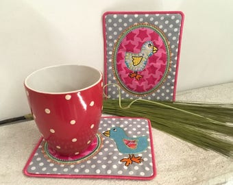 May include: Two gray and pink fabric coasters with a white polka dot pattern. One coaster features a pink oval with a yellow bird and stars. The other coaster features a blue bird with orange feet.