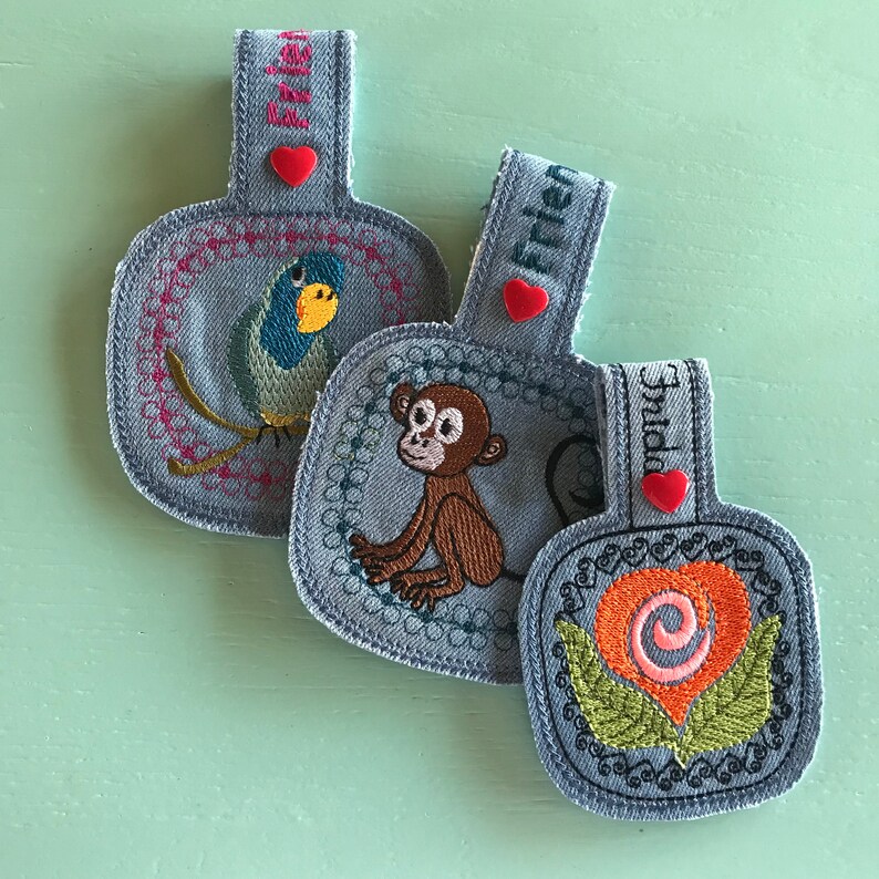 May include: Three blue denim fabric keychains with embroidered designs. The first keychain features a blue and yellow bird, the second keychain features a brown monkey, and the third keychain features a pink and orange flower.