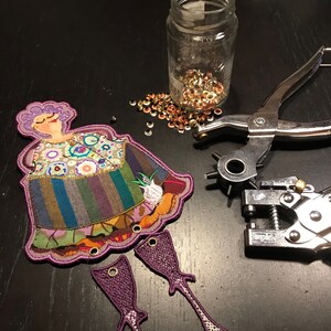 Peut inclure: Une poupée en tissu violet avec une robe colorée et un motif floral. La poupée manque de bras et de jambes, qui sont présentés séparément. La poupée est sur une surface sombre avec un pot d'œillets métalliques et deux outils de poinçonnage métalliques.