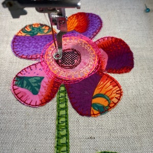 Puede incluir: Primer plano de una aguja de m&aacute;quina de coser cosiendo una flor de tela rosa y morada con hojas verdes. La flor tiene un centro rosa con un dise&ntilde;o circular.